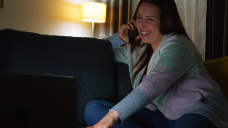 Smiling-Woman-Sitting-On-Sofa-At-Home-At-Night-Talking-On-Mobile-Phone-And-Watching-Movie-Or-Show-On-Laptop-9